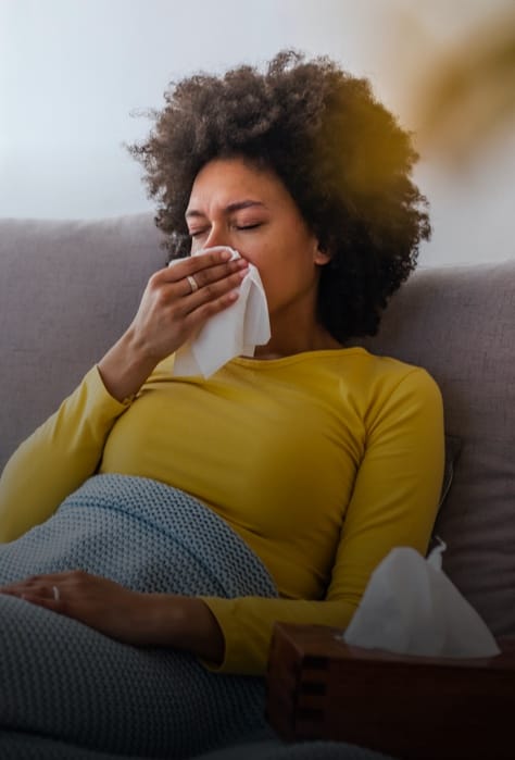 Woman sitting on couch with blanket blowing her nose with tissues